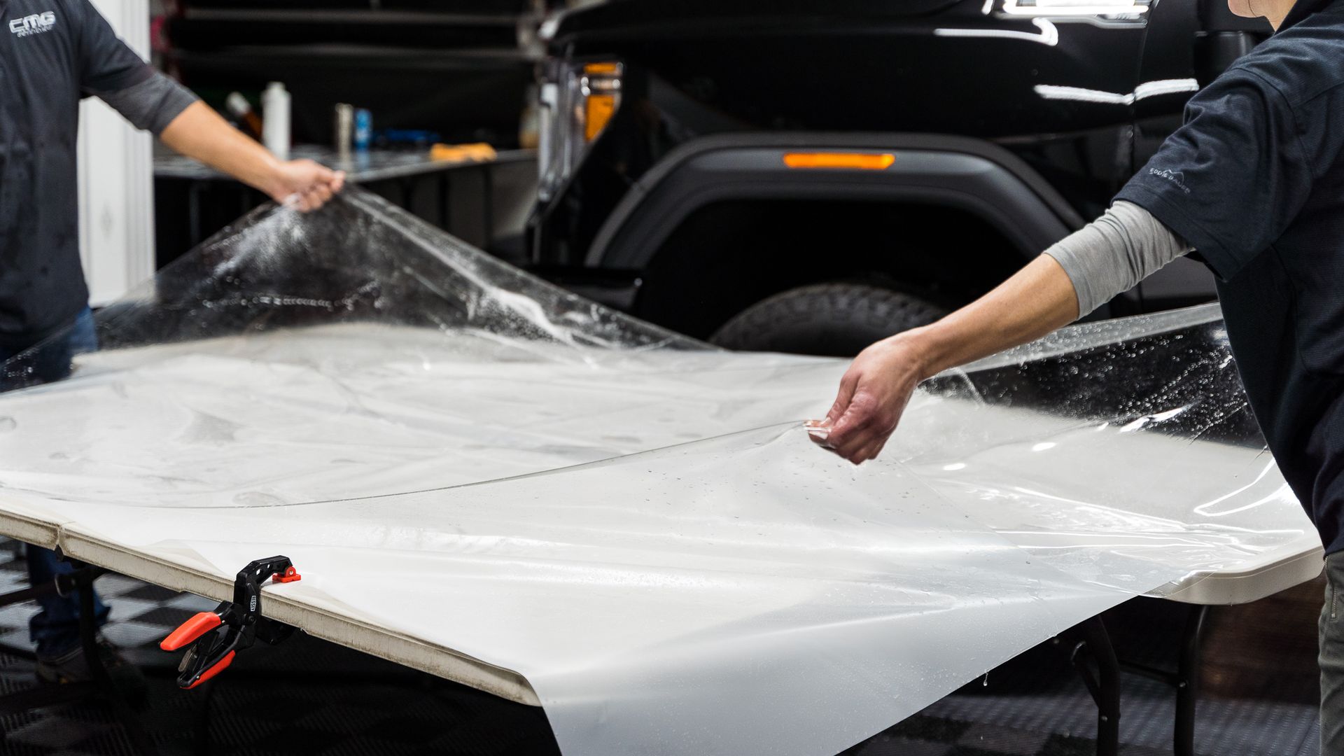 Two people applying clear film to a table near a black vehicle.
