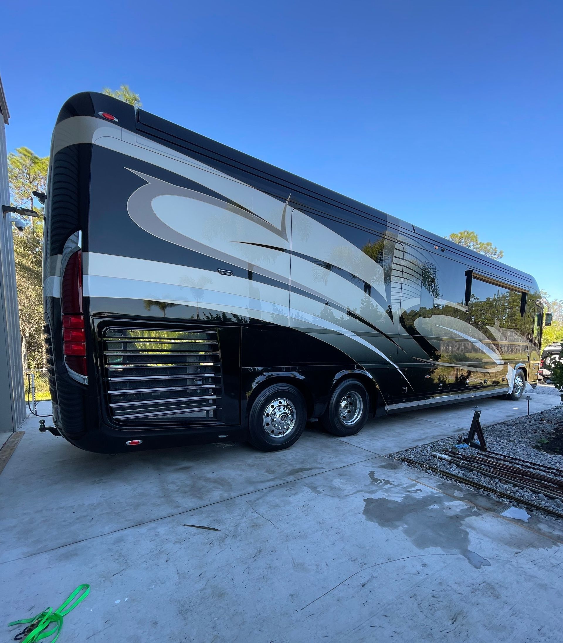 Large, black and silver luxury RV parked on a concrete surface, with trees in the background under a blue sky.