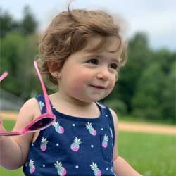 Smiling child in a blue pineapple dress holding pink sunglasses outdoors