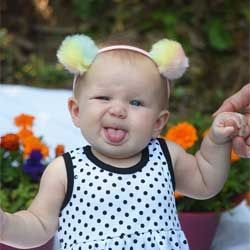Toddler winking and sticking out tongue, wearing polka-dot dress and fuzzy headband with pom-poms