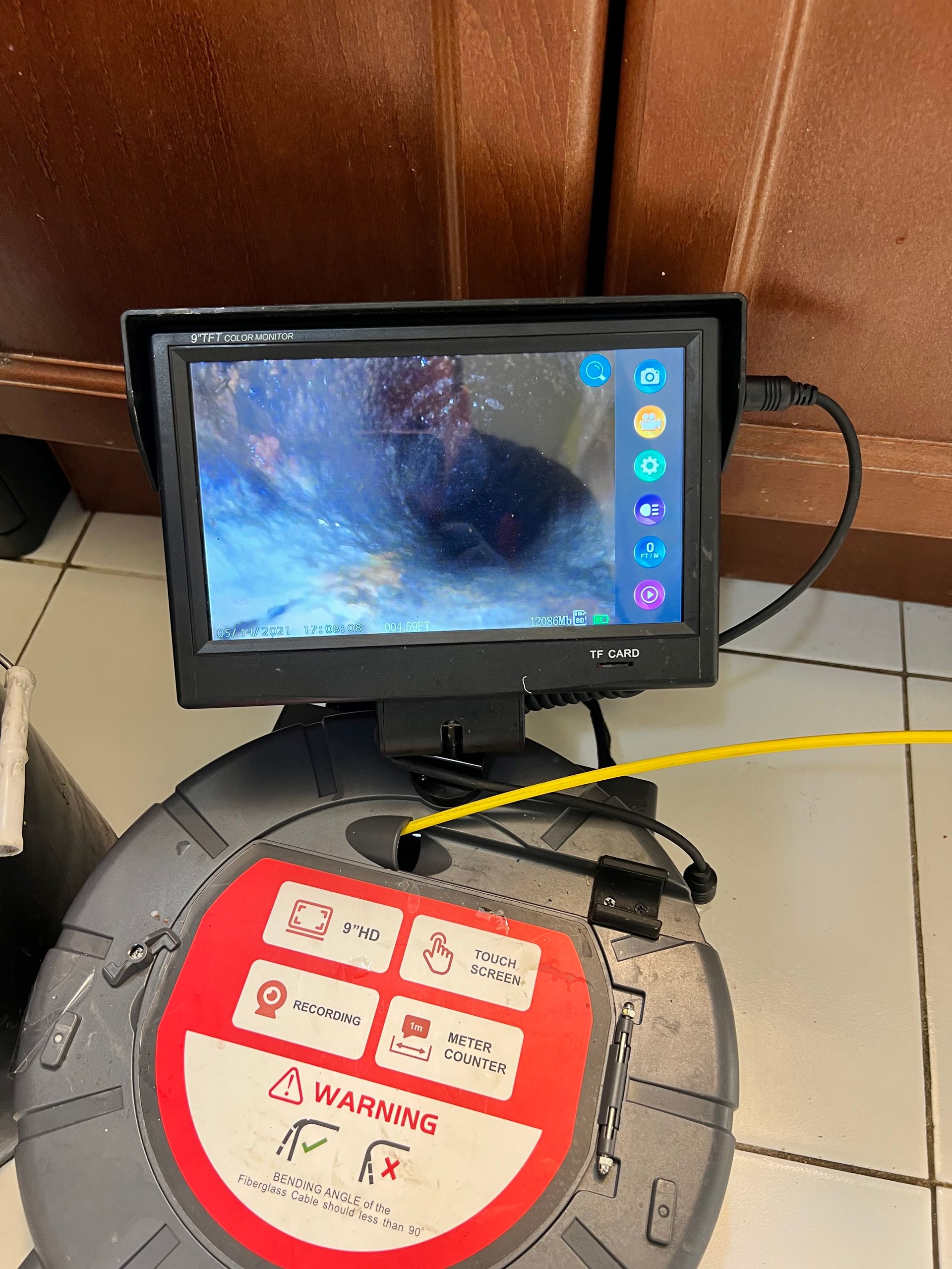 A plumbing camera screen displays a pipe interior. A reel with controls and a warning sticker sits below.