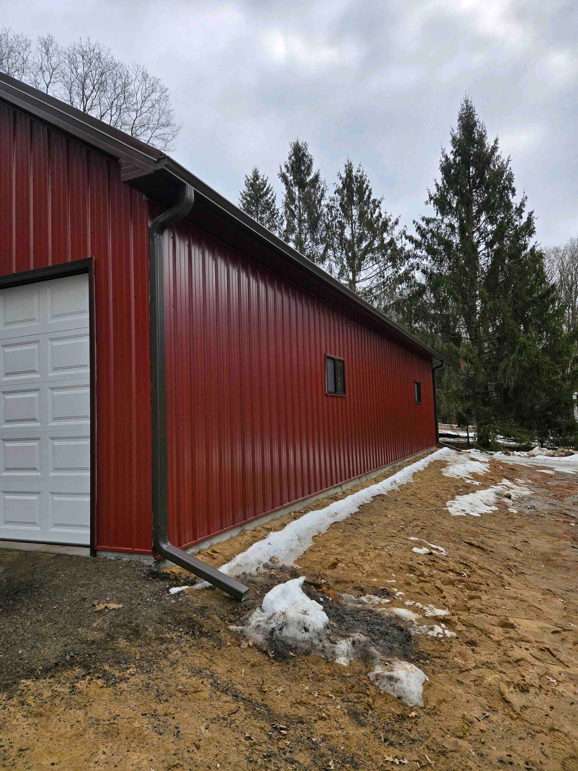 Red barn with white garage door beside a snowy gravel path under cloudy skies.