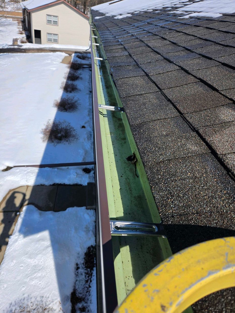 Snow-covered roof edge beside shingles, with exposed green gutter and a yellow safety bucket in view