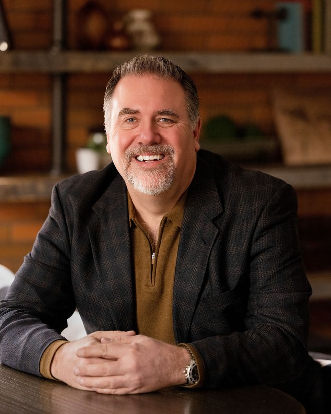 Smiling man in a dark blazer sits at a table in a warmly lit room.
