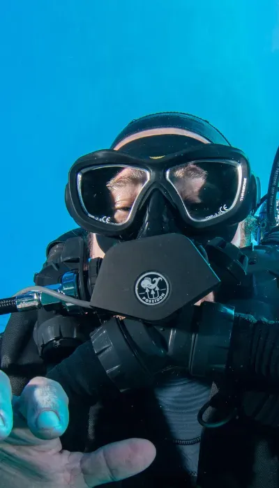 Scuba diver in black mask and gear underwater, holding the camera close against a blue background