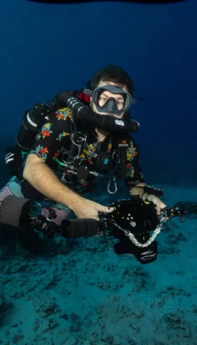 Scuba diver in black gear kneeling on the ocean floor, holding equipment against a blue underwater background
