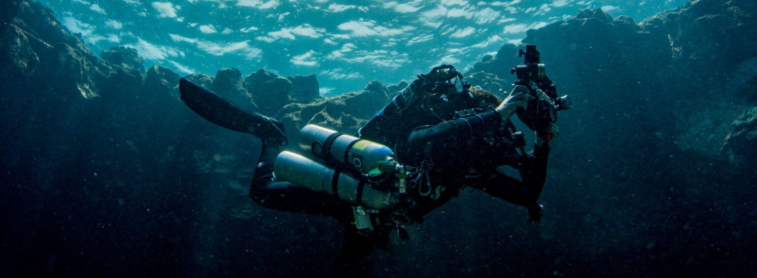Two scuba divers underwater, swimming through a blue ocean with bubbles and gear.