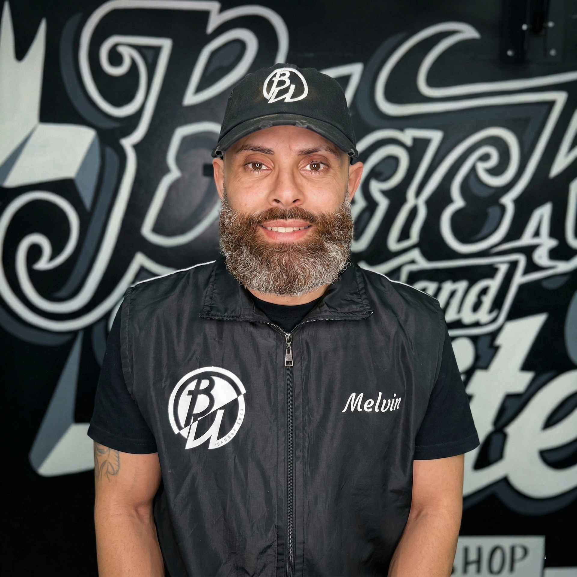 Man with beard and hat in front of a shop sign. Wearing a vest with logo and the name 