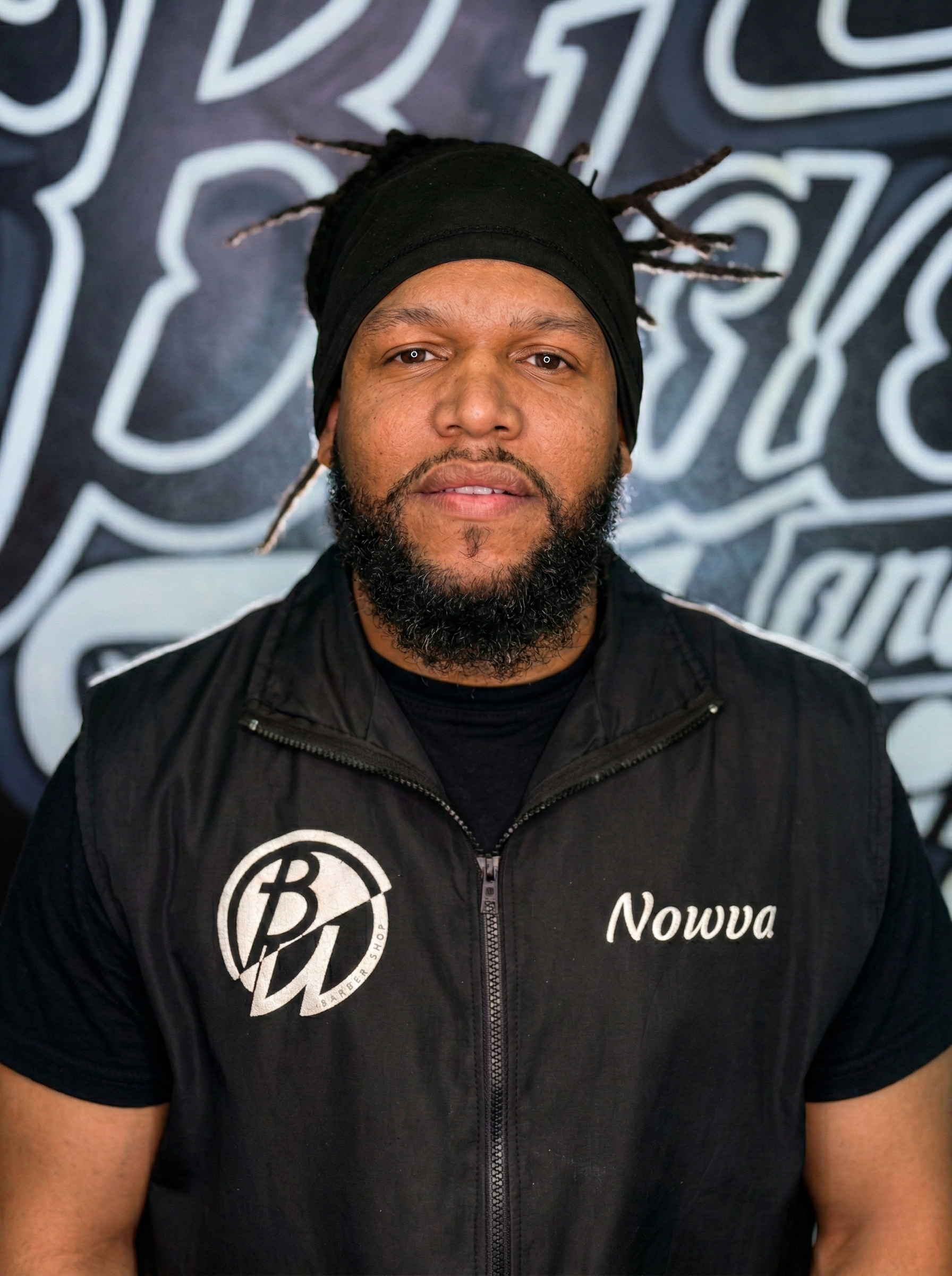 Man with a black beard wearing a black vest and beanie, standing in front of a logo.