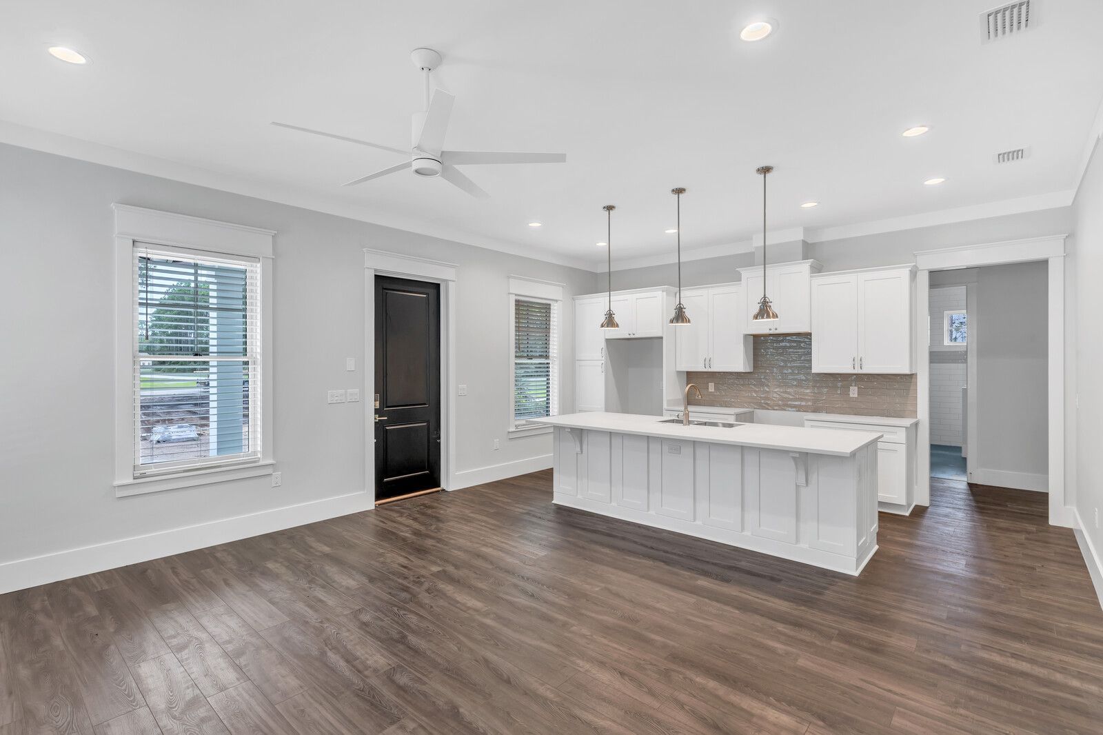 A living room with hardwood floors and a kitchen with white cabinets and a ceiling fan.