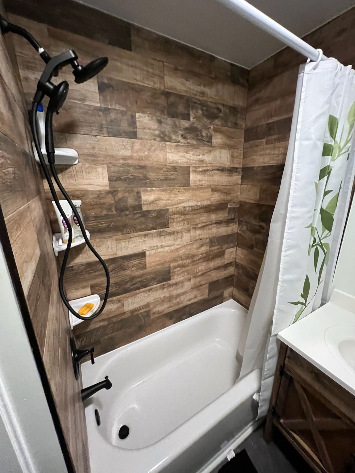 Shower with wood-look tile walls, white bathtub, black shower fixtures, and a green leaf shower curtain.