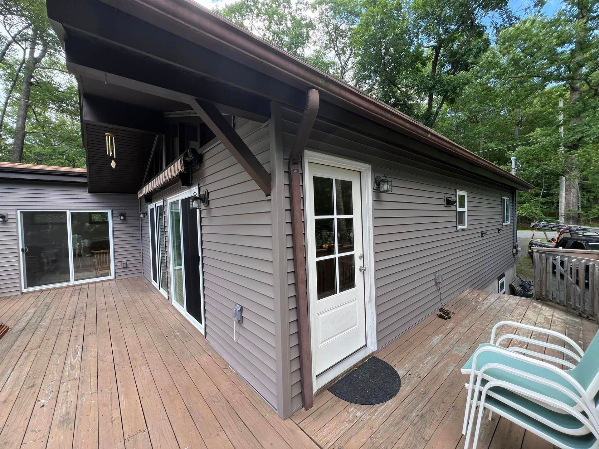 Exterior of a gray-sided cabin with a white door on a wooden deck surrounded by trees.