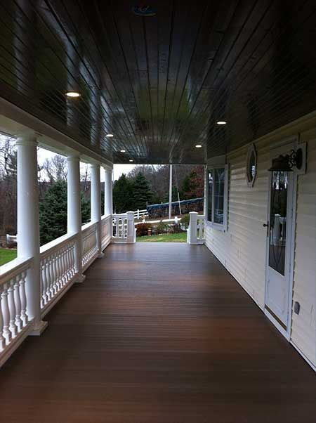 Covered porch with white columns, railing, brown deck and ceiling, and a white door.