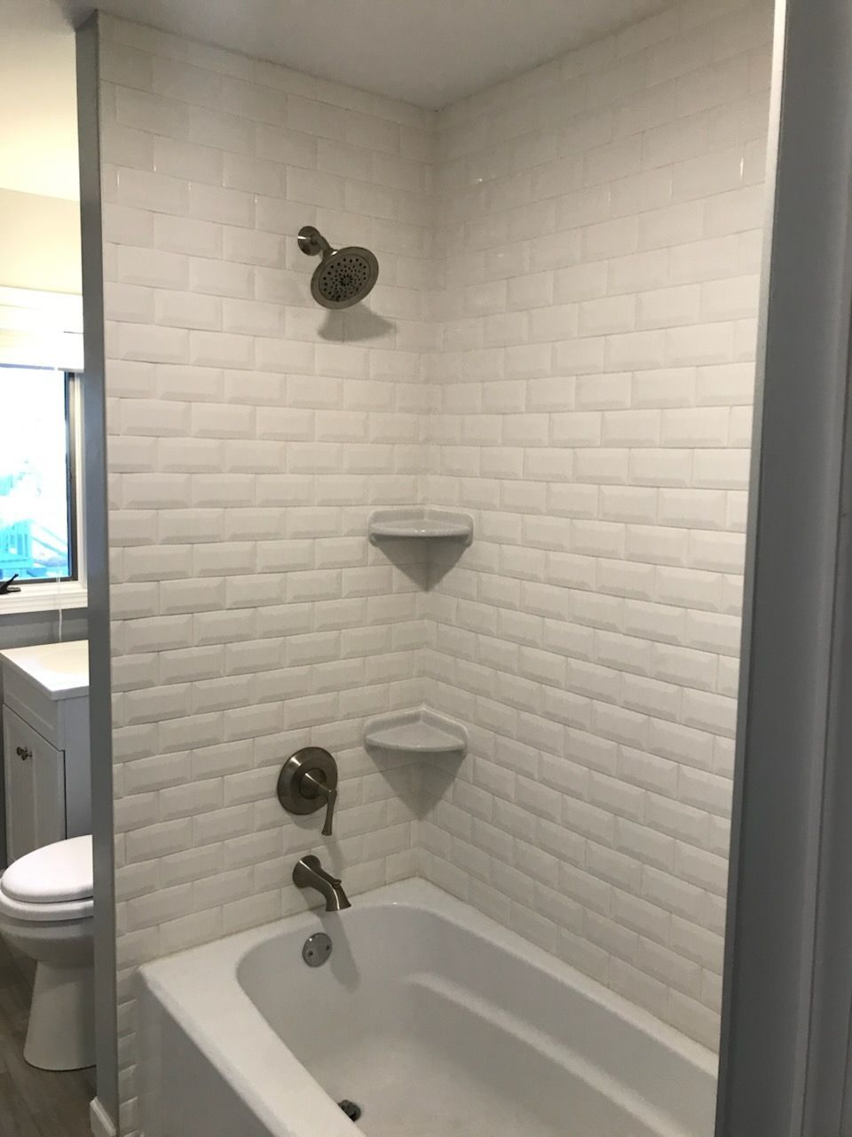 White-tiled shower with a tub, showerhead, two corner shelves, and faucet. A toilet and vanity are visible on the left.