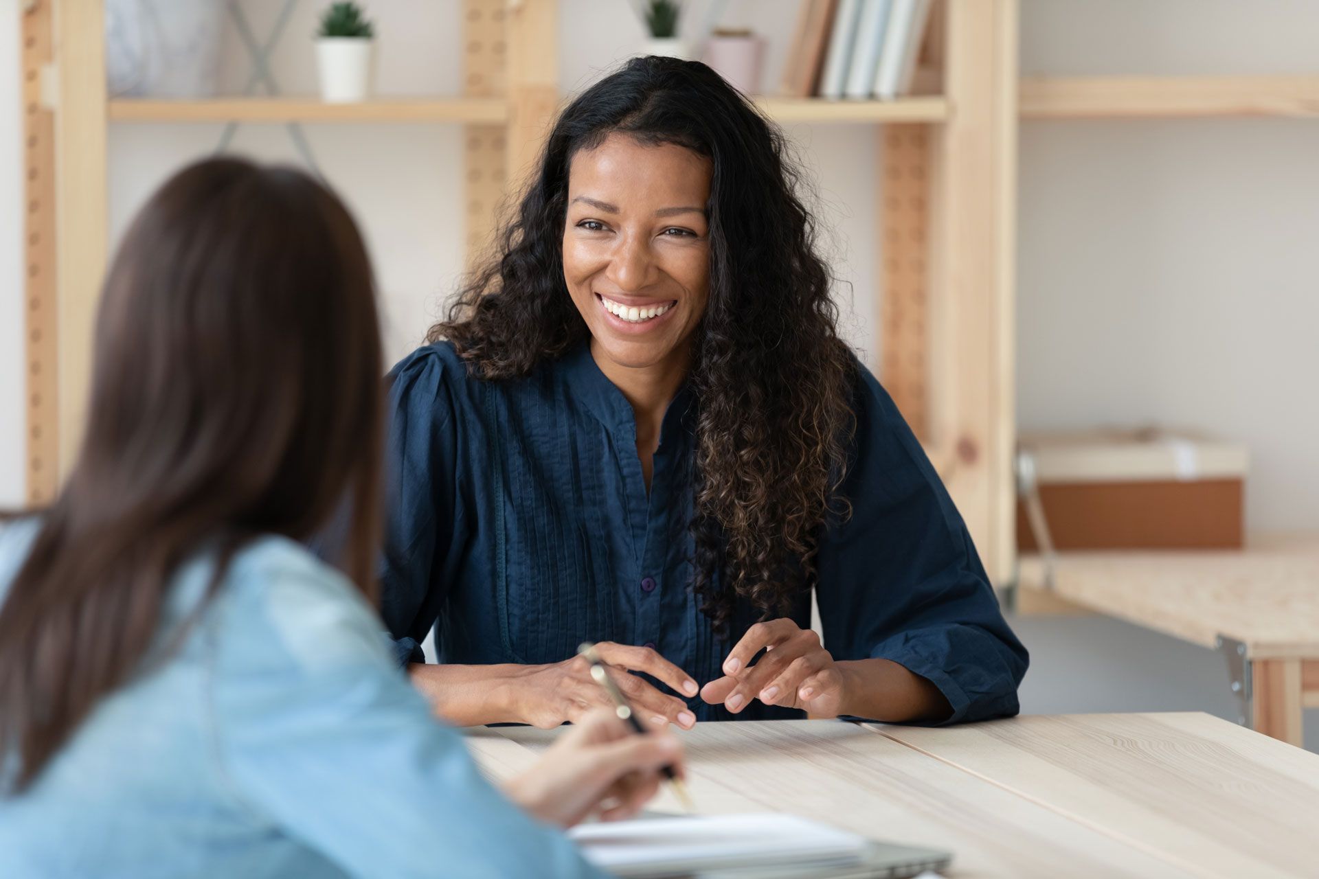 Woman smiles while talking to another person at a desk. Wooden shelves are in the background.