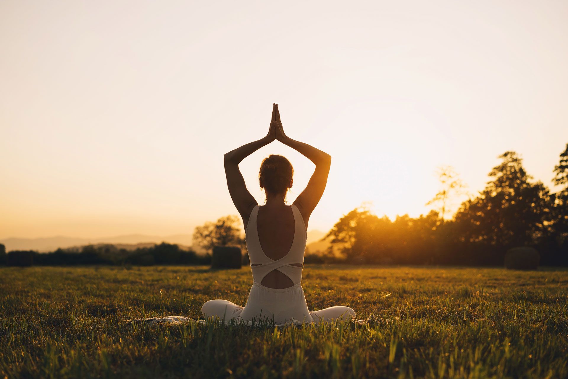 Person in white outfit meditating outdoors with hands together, sunset in background.