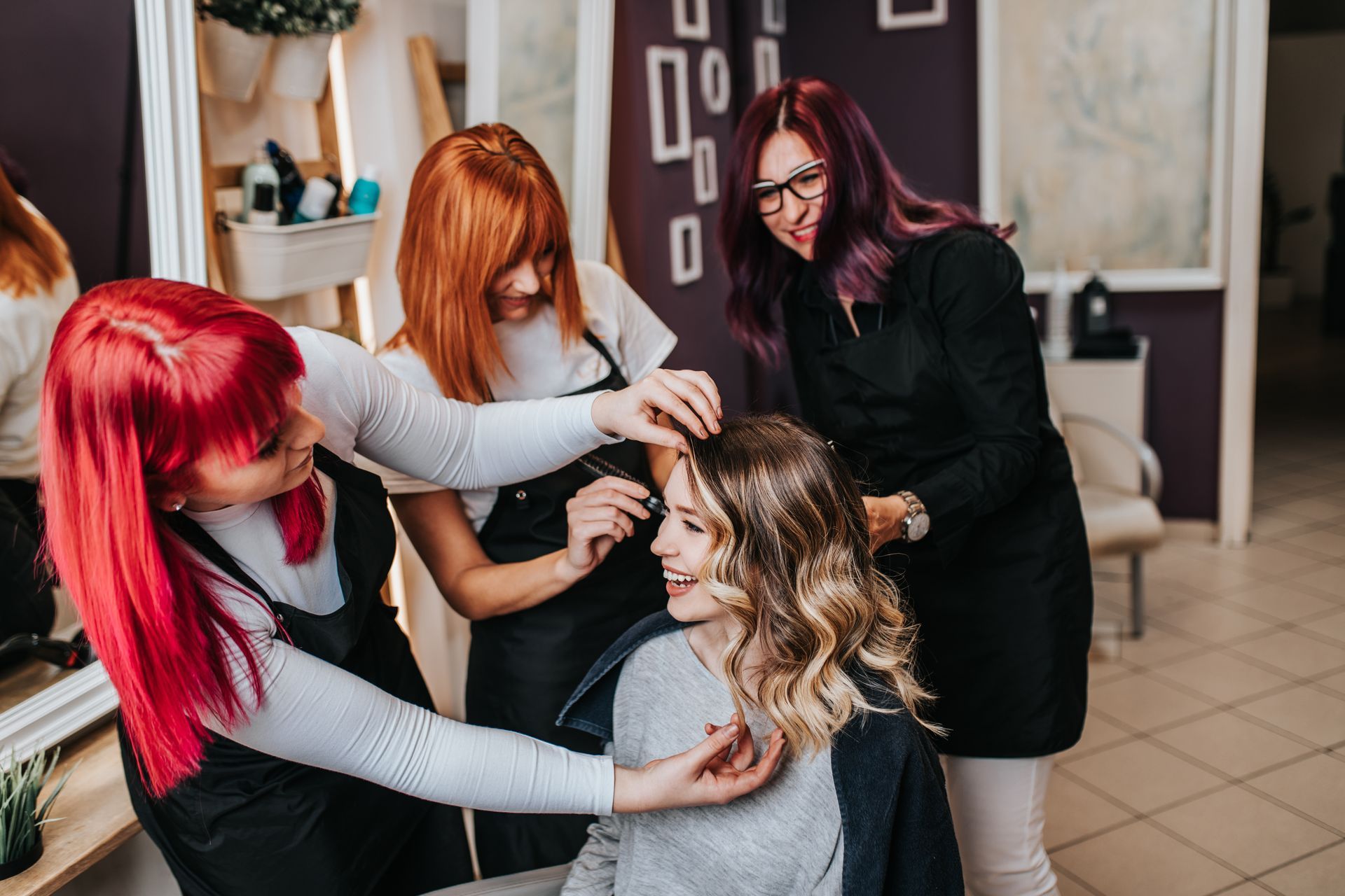 Four people in a salon: three stylists working on a client's wavy, layered hair. Two stylists have red hair.