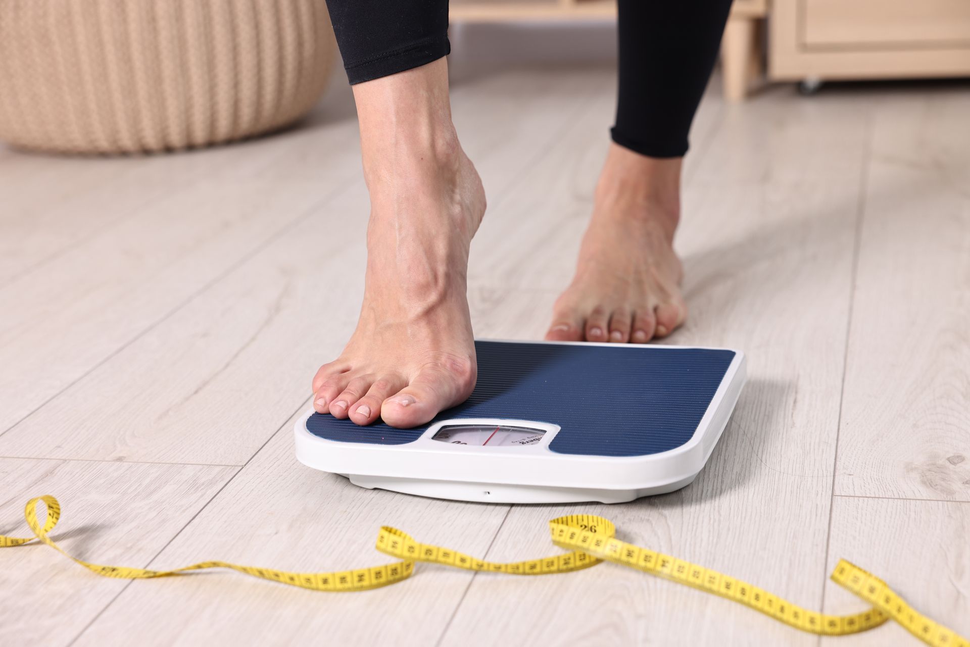 Person's bare foot stepping onto a blue and white scale on a wooden floor, with a yellow measuring tape nearby.