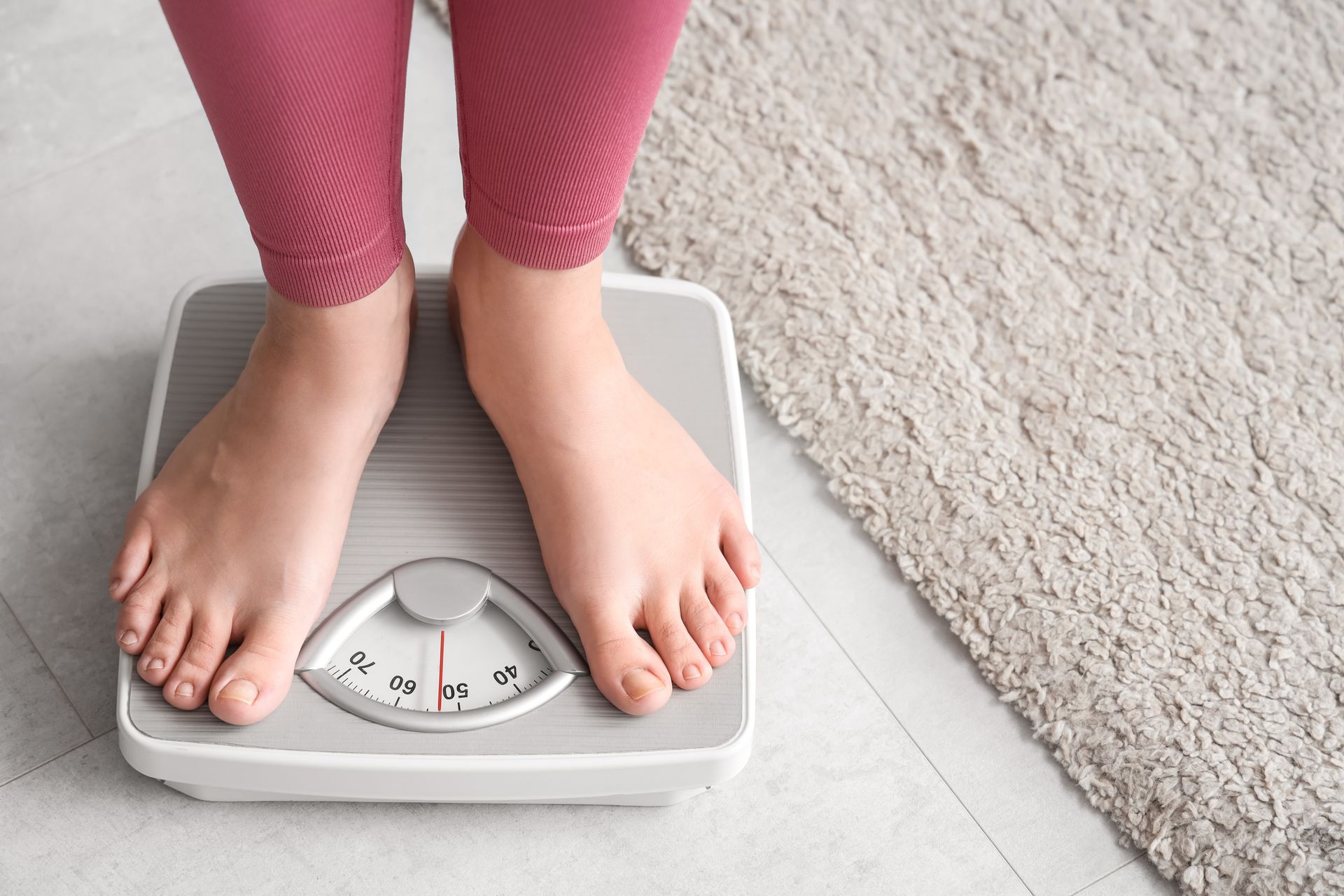 Person's feet on a scale, wearing pink leggings. A beige rug is to the right.
