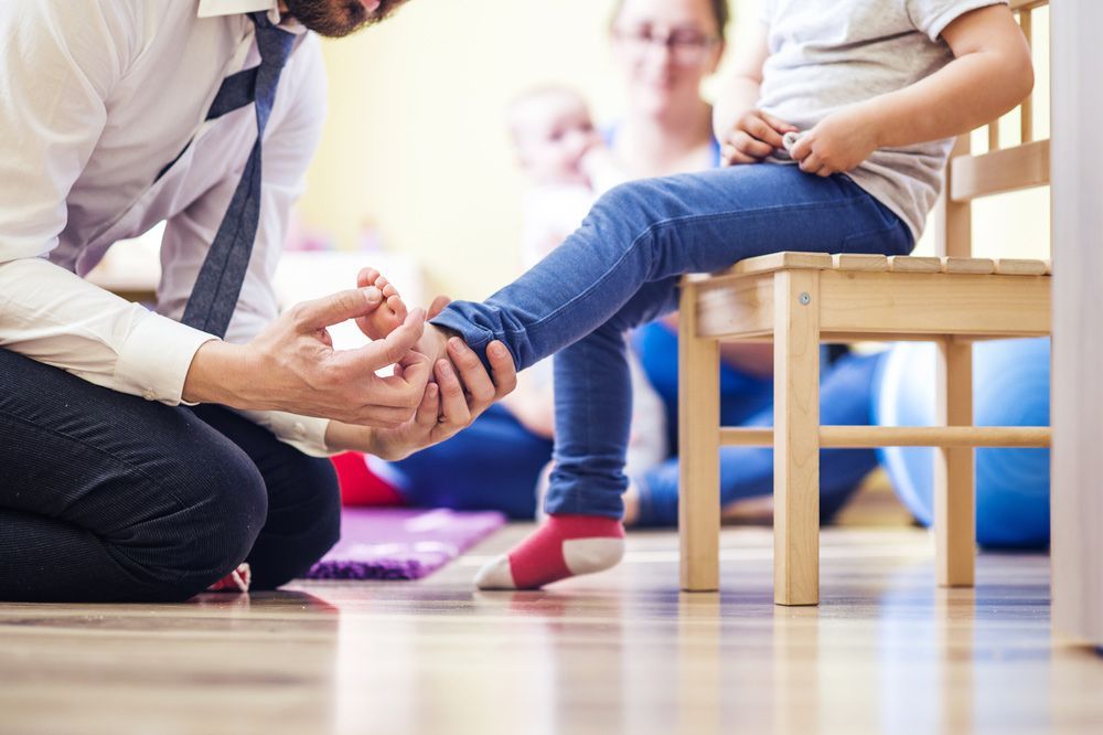 A Man Is Kneeling Down To Help A Little Girl Put On Her Shoes — Sandy Gugenberger Podiatrist in Port Macquarie, NSW