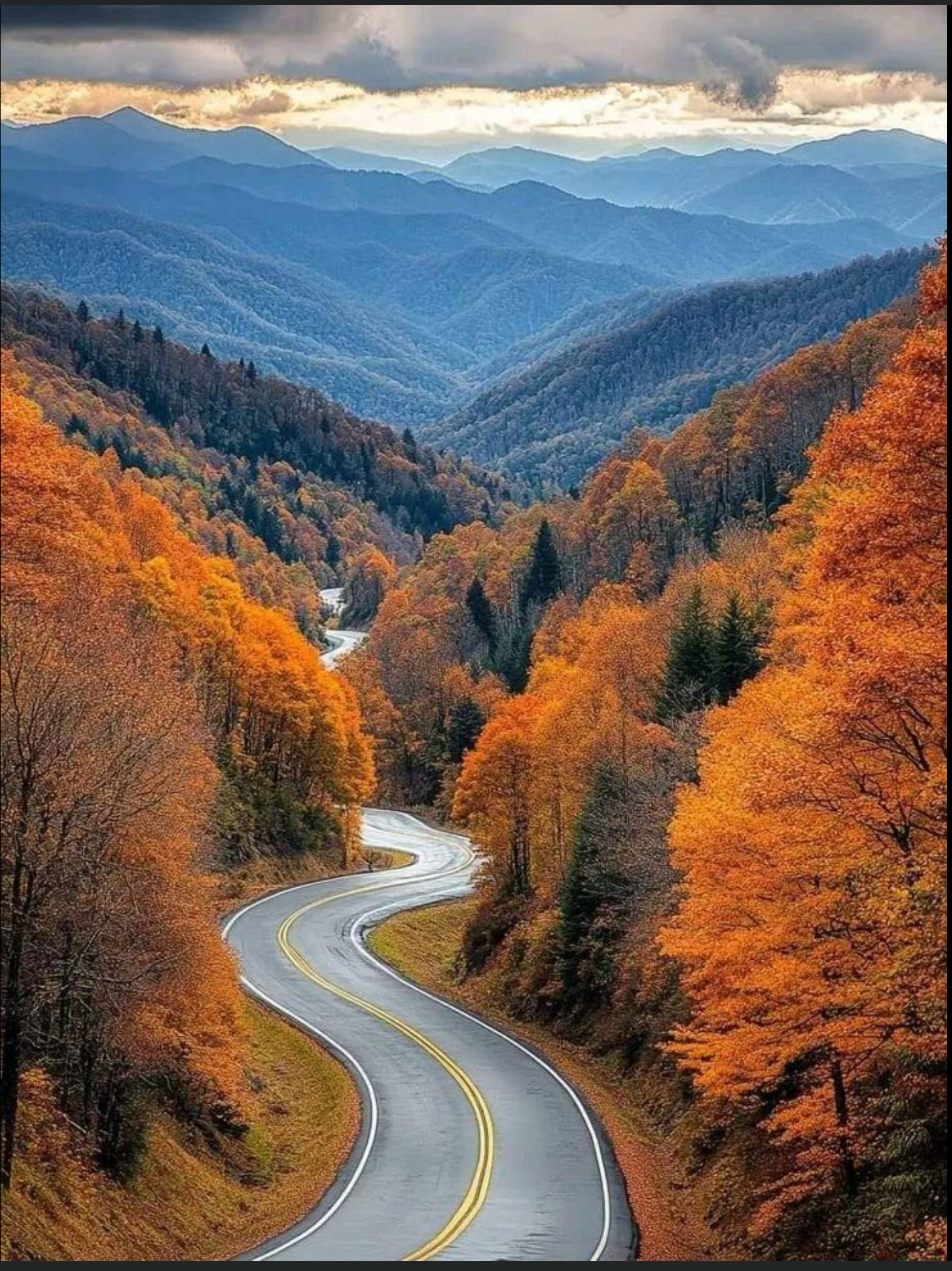 Winding mountain road through orange autumn forest, with blue hills and cloudy sky beyond