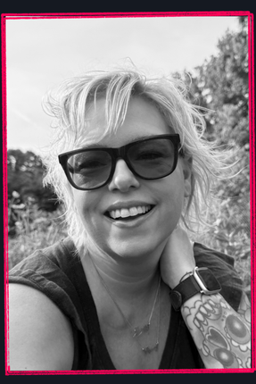 Smiling person wearing sunglasses, outdoors, with windblown hair and a tattooed arm.