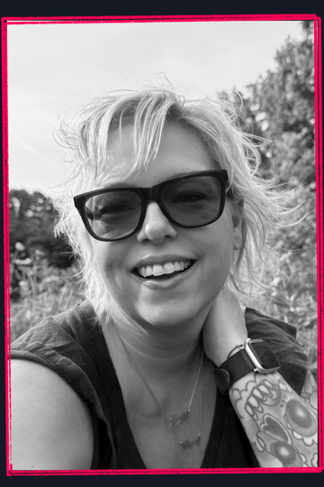 Smiling person wearing sunglasses, outdoors, with windblown hair and a tattooed arm.