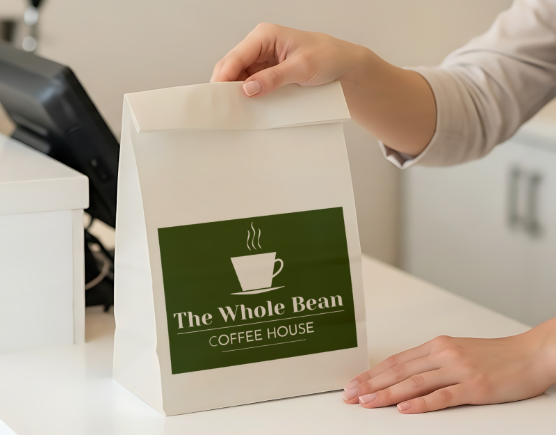 Hand placing a white coffee bag with green “The Whole Bean Coffee House” label on a counter