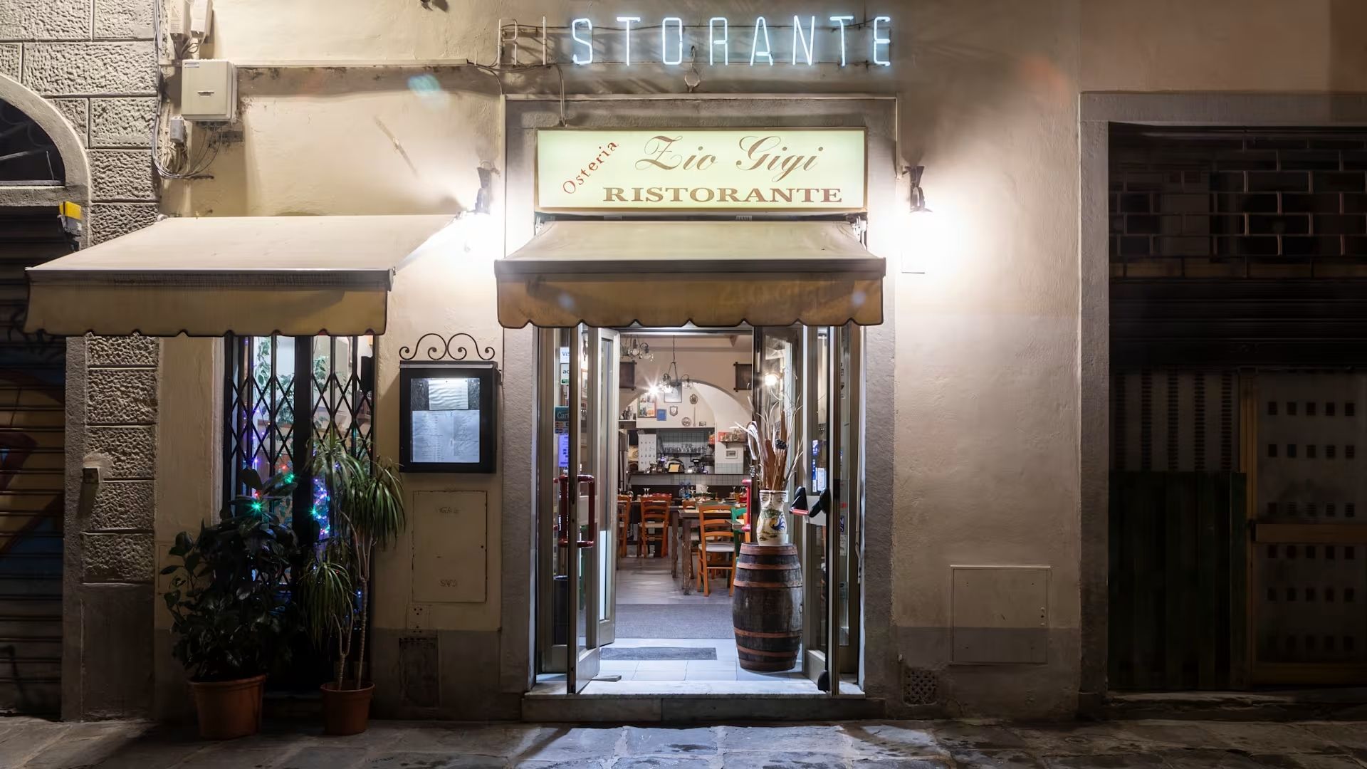 L'ingresso del ristorante al crepuscolo, con un'insegna illuminata, la porta aperta e i tavoli visibili all'interno.