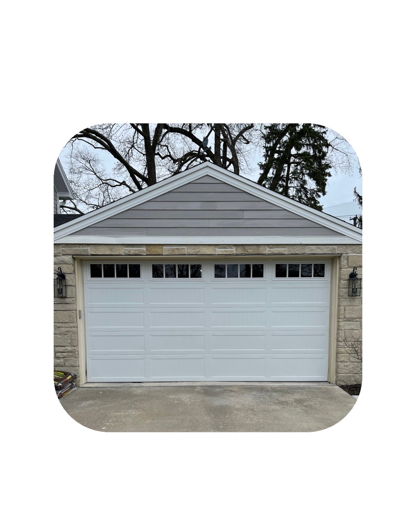 White garage door with gray roof and stone siding, trees in background.