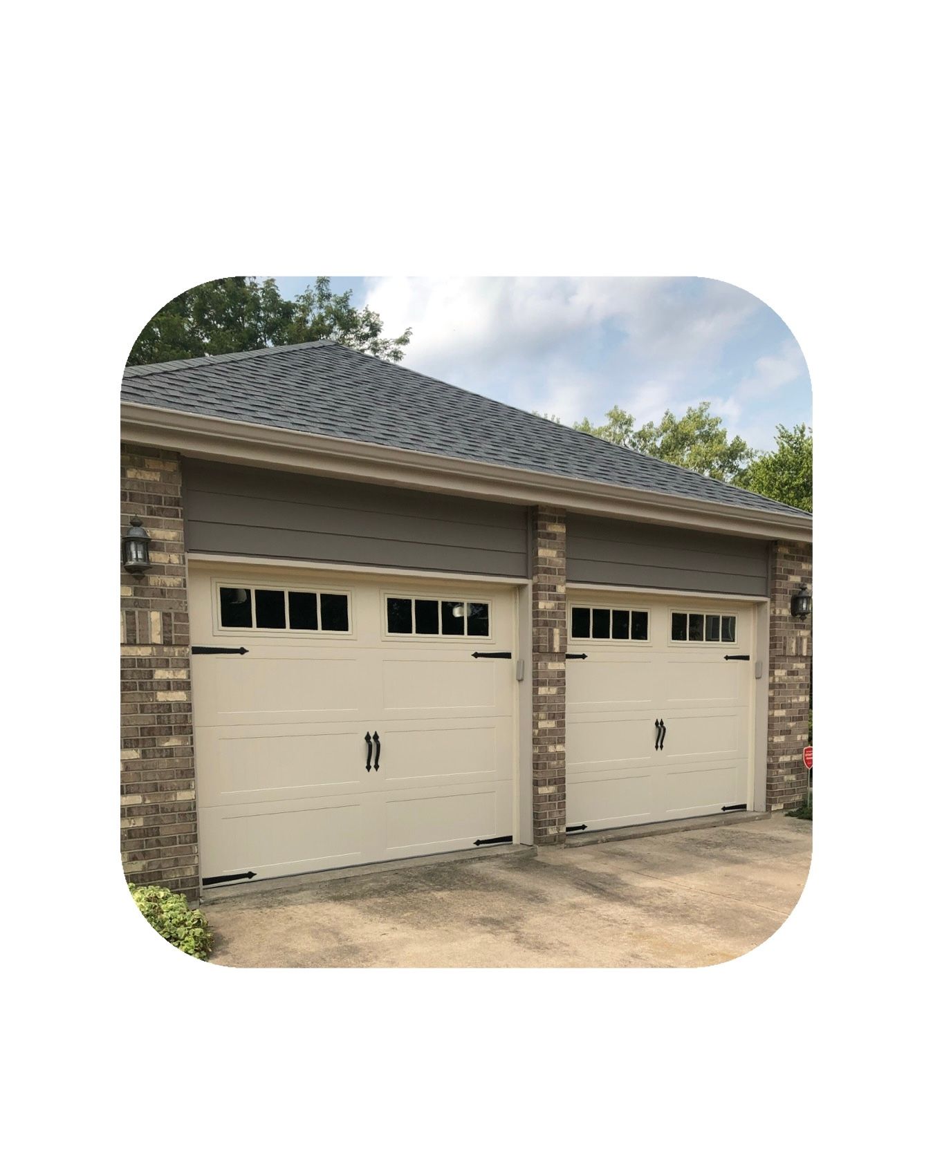 Two-car garage with tan doors, brick accents, and a dark shingled roof under a partly cloudy sky.