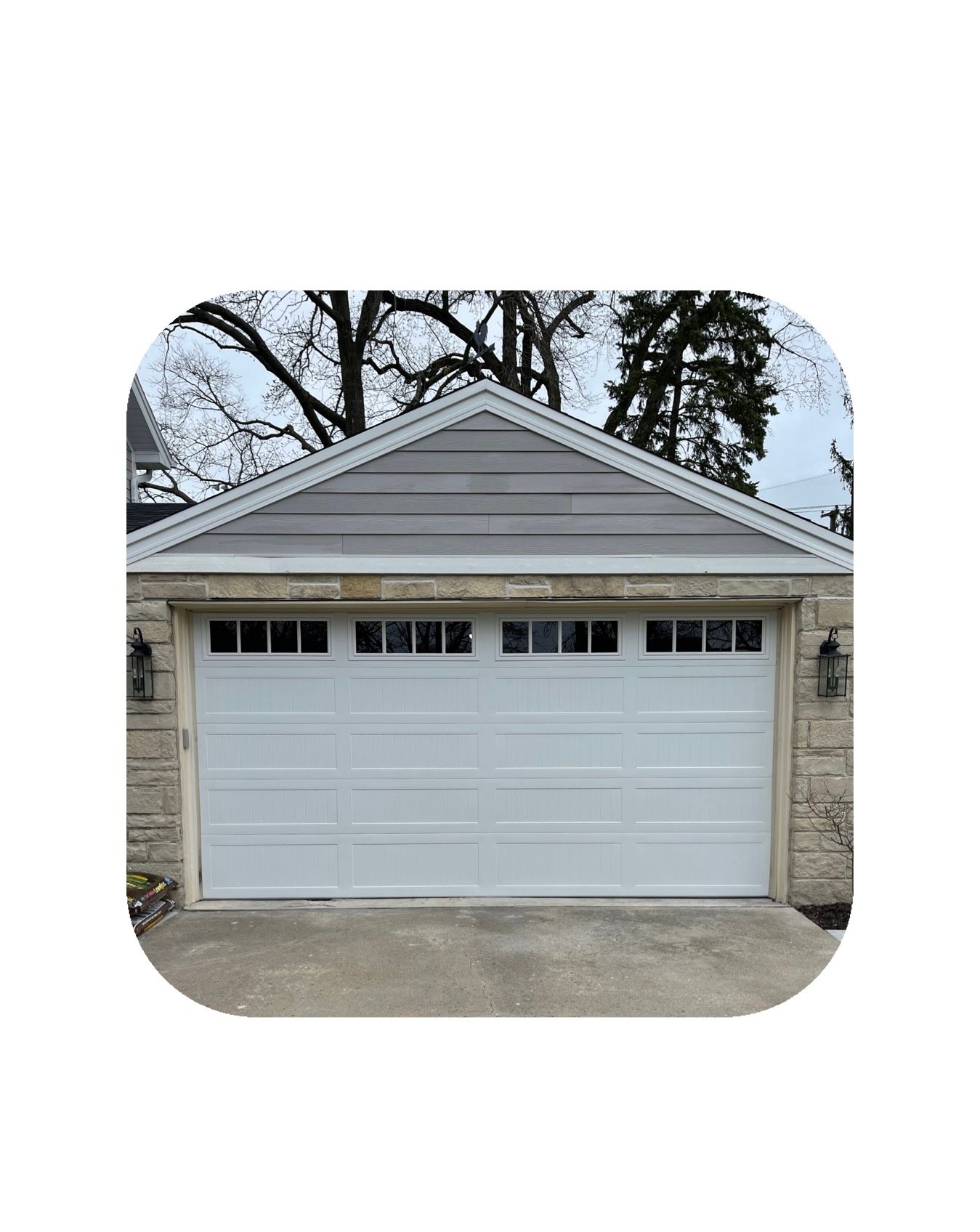 White garage door with gray roof, set in a stone facade, against a backdrop of bare trees.