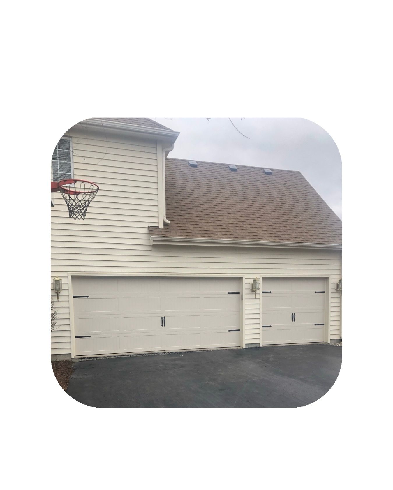 Beige two-car garage with a basketball hoop on the side, asphalt driveway, and a brown shingled roof.