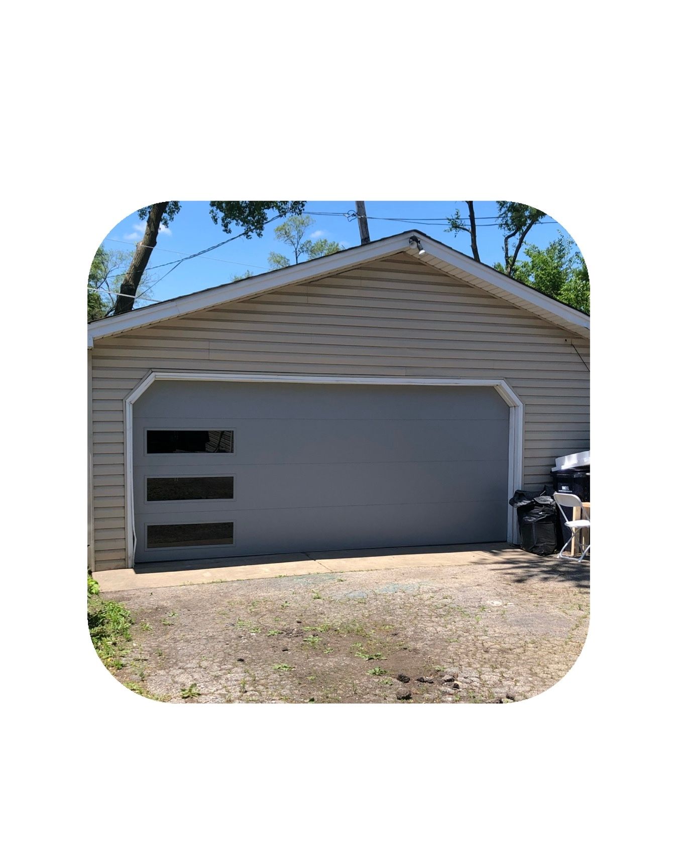 Gray garage door with three horizontal window panels. Beige siding, gravel driveway, and trees.