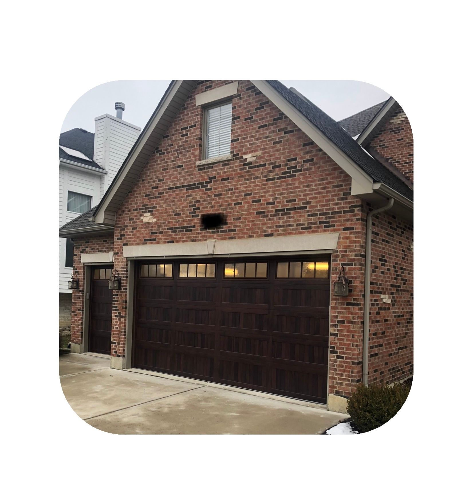 Brown garage doors on a brick house with a small window above. Snow on the ground.