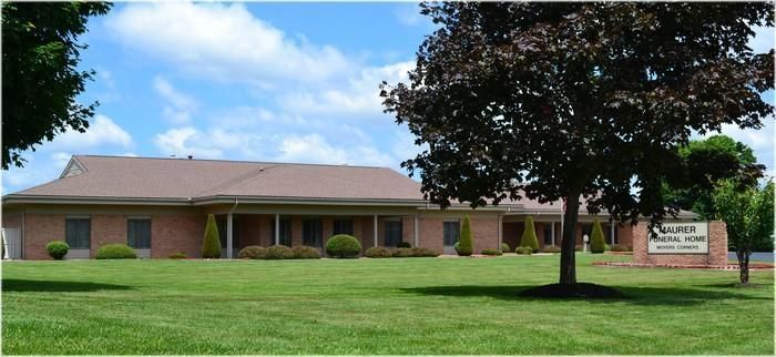 Single-story brick building with a brown roof and a sign in a grassy front yard under a blue sky with scattered clouds.