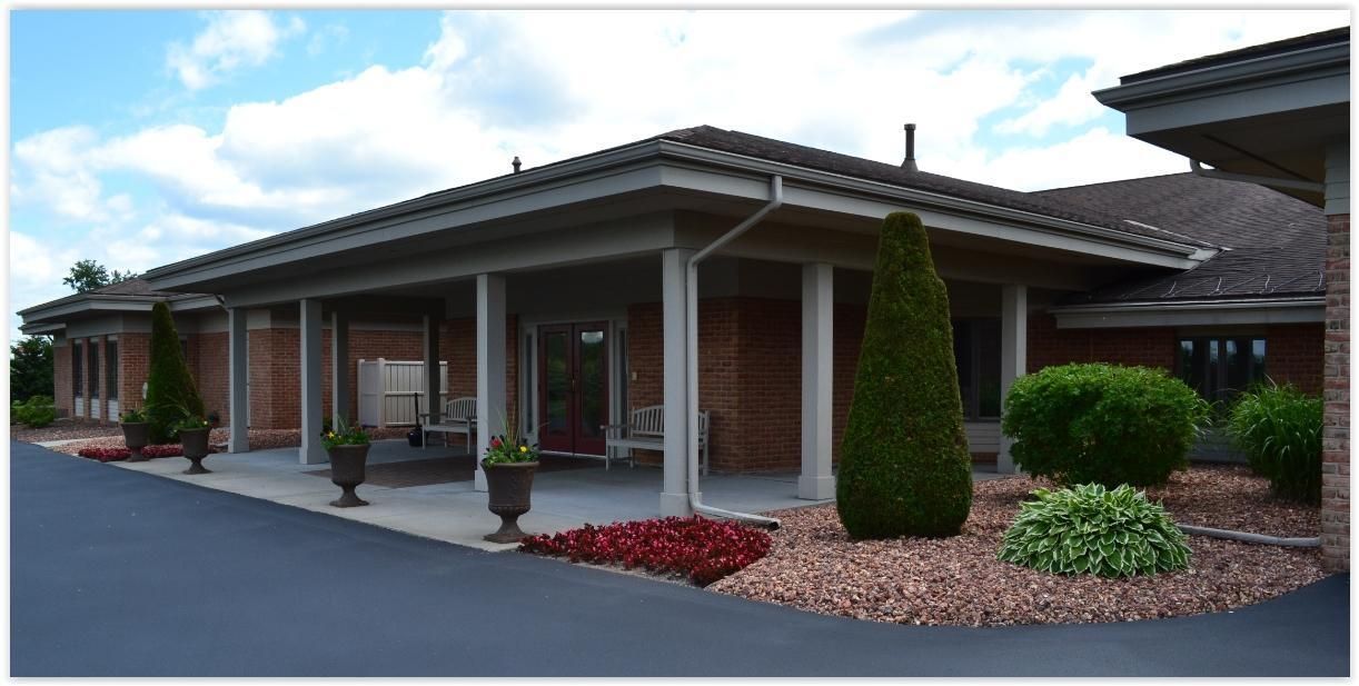 A single-story brick building with a covered entrance portico, featuring landscaped gardens with bushes and flowers.