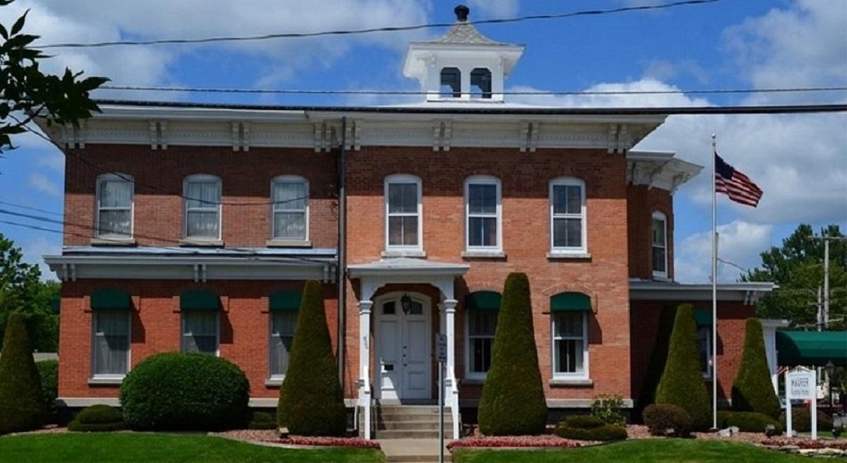 A two-story brick building with a central cupola, a front entrance porch, and an American flag on a pole to the right.