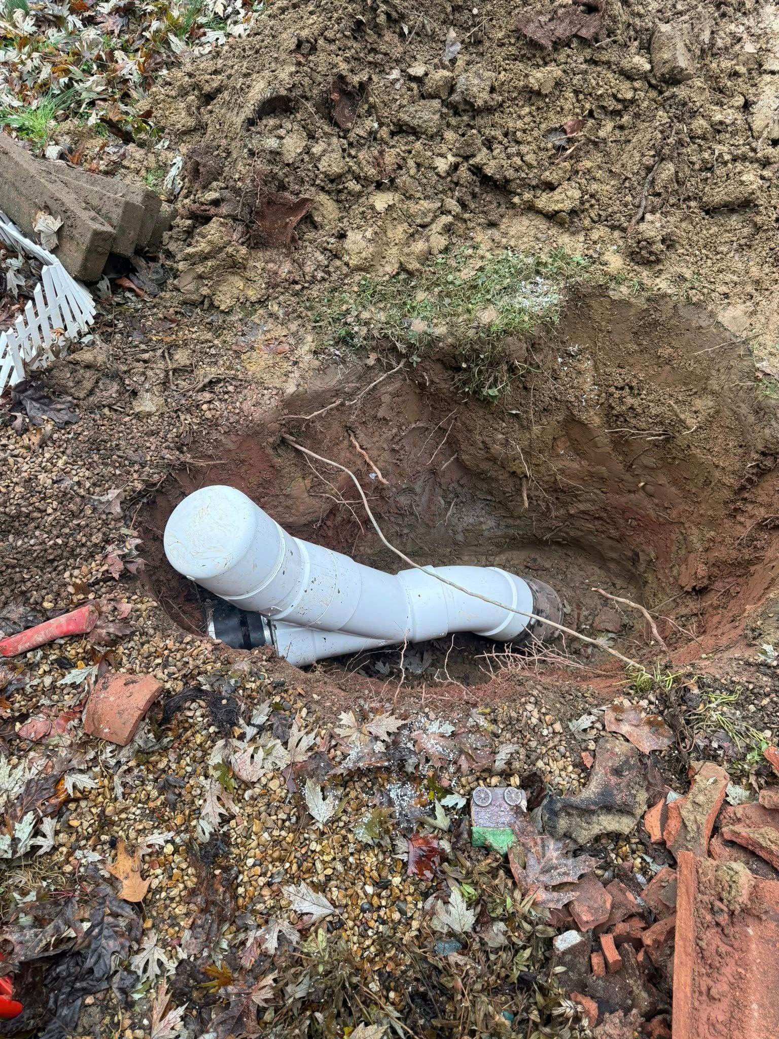 A white PVC pipe assembly with a capped end is buried in a hole in the ground with loose dirt and debris nearby.