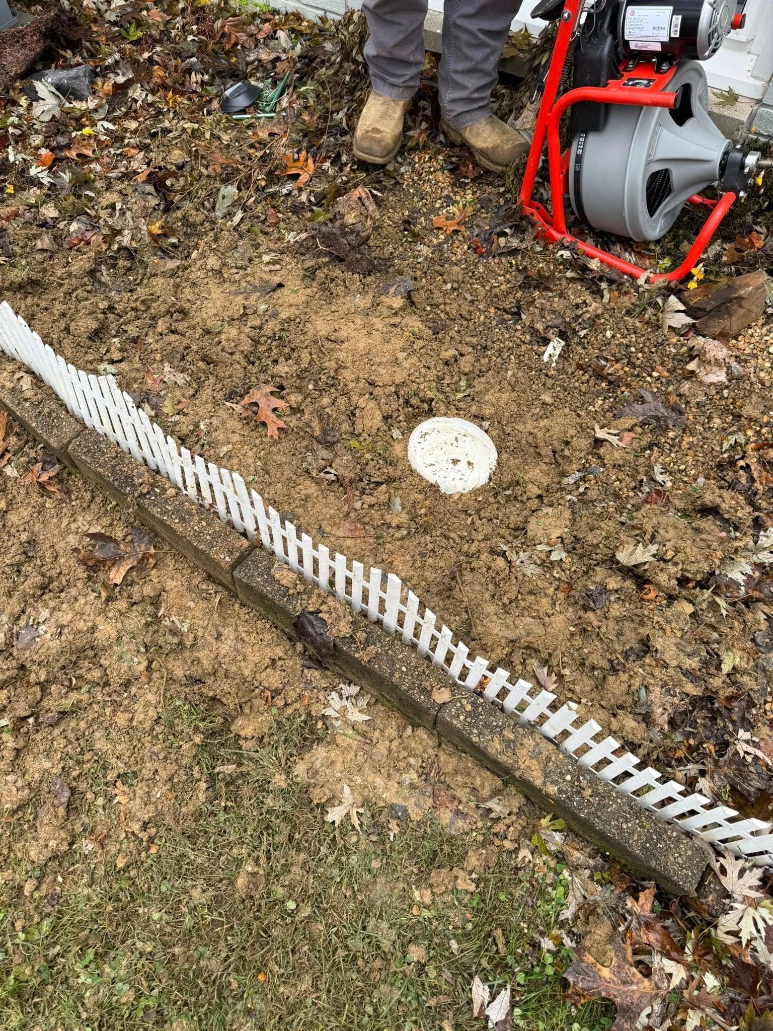 A technician stands by a sewer auger on the ground near a white access pipe and decorative white garden fence.