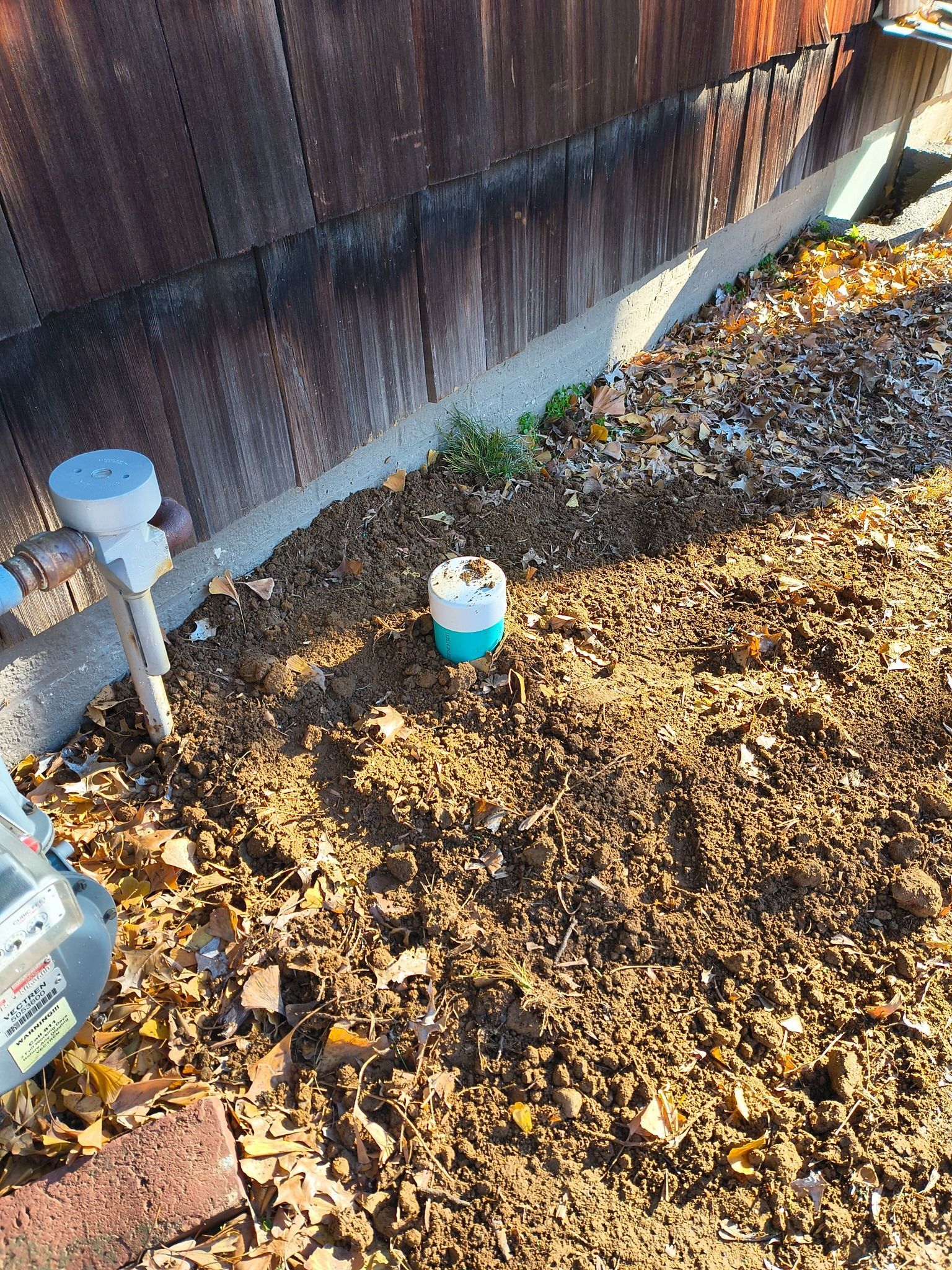 A PVC pipe cleanout cap protrudes from soil and fallen leaves next to a building's foundation and a gas meter.