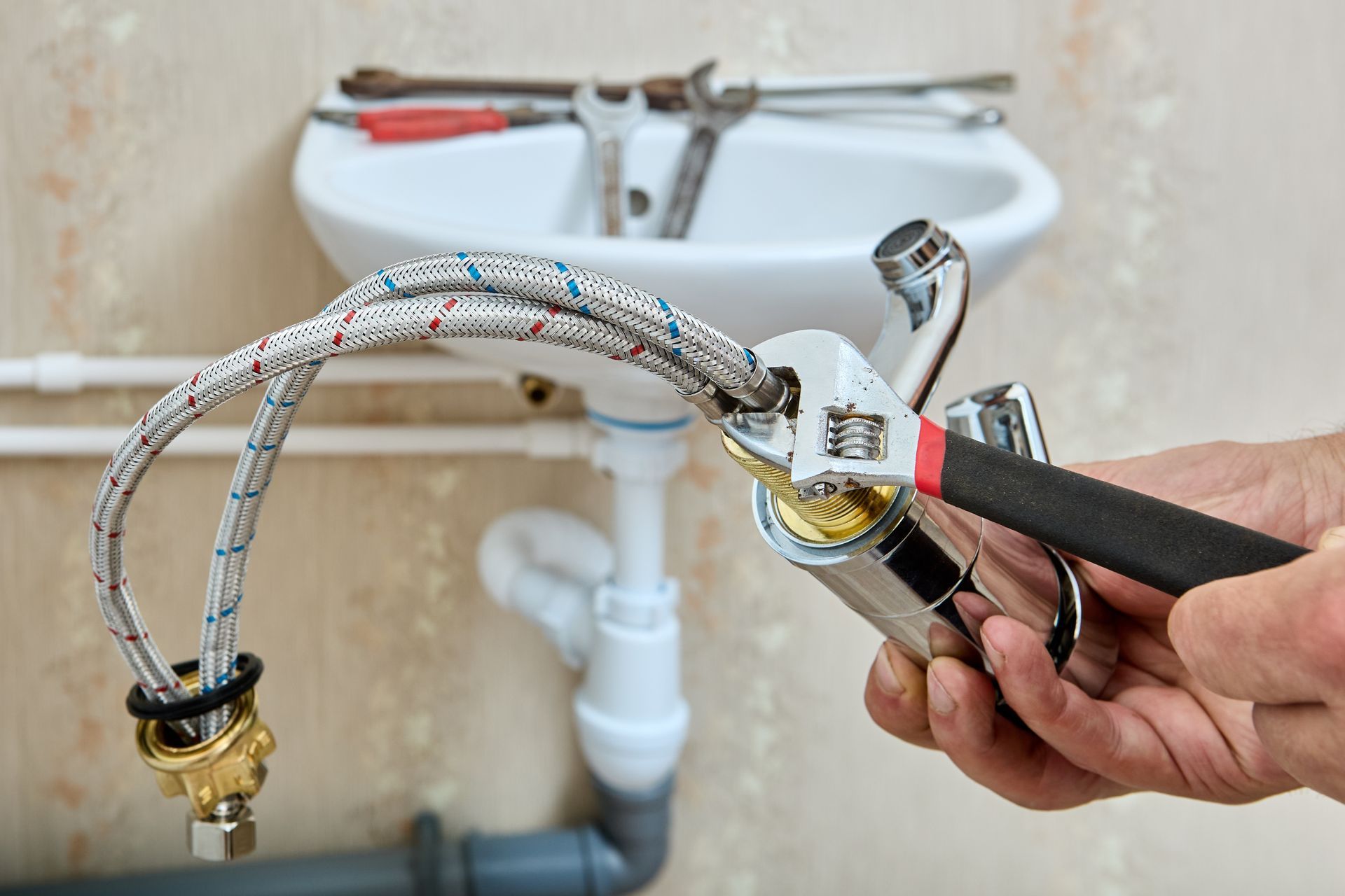 Hands using an adjustable wrench to tighten a metal hose onto a faucet in front of a white sink.