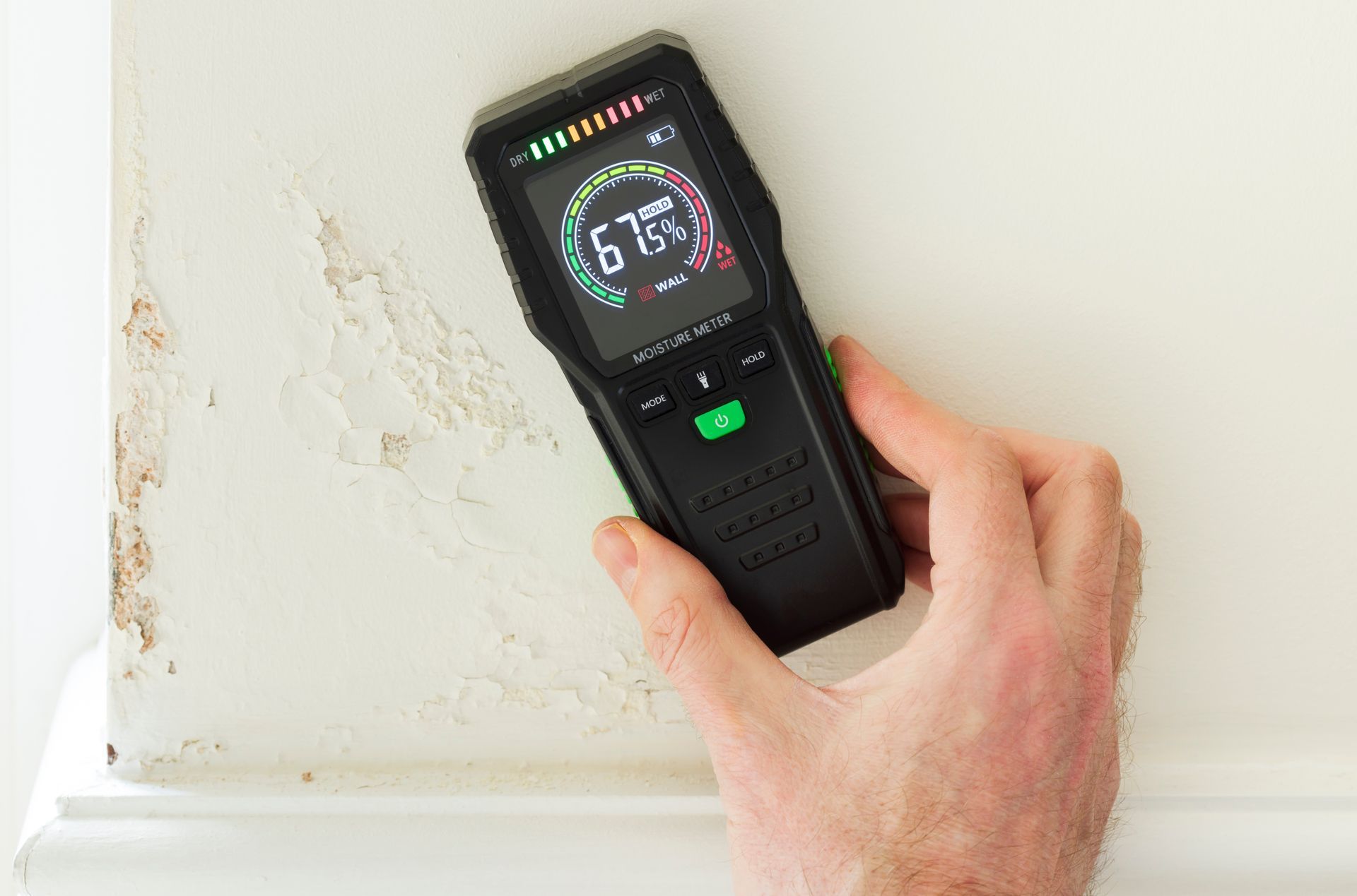 A hand holds a moisture meter against a wall with visible water damage, showing a reading of 67%.