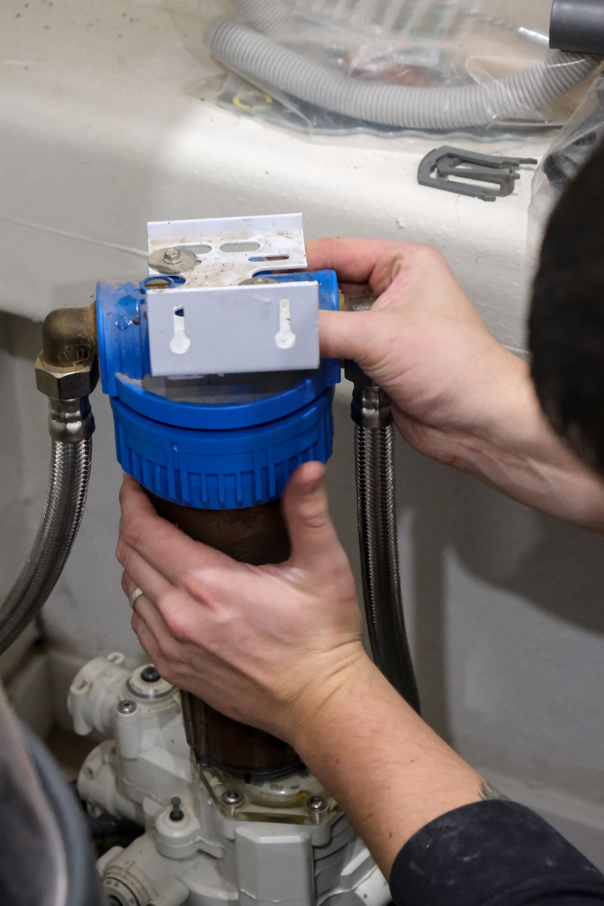 Hands holding a blue water filter housing while performing maintenance on plumbing equipment.