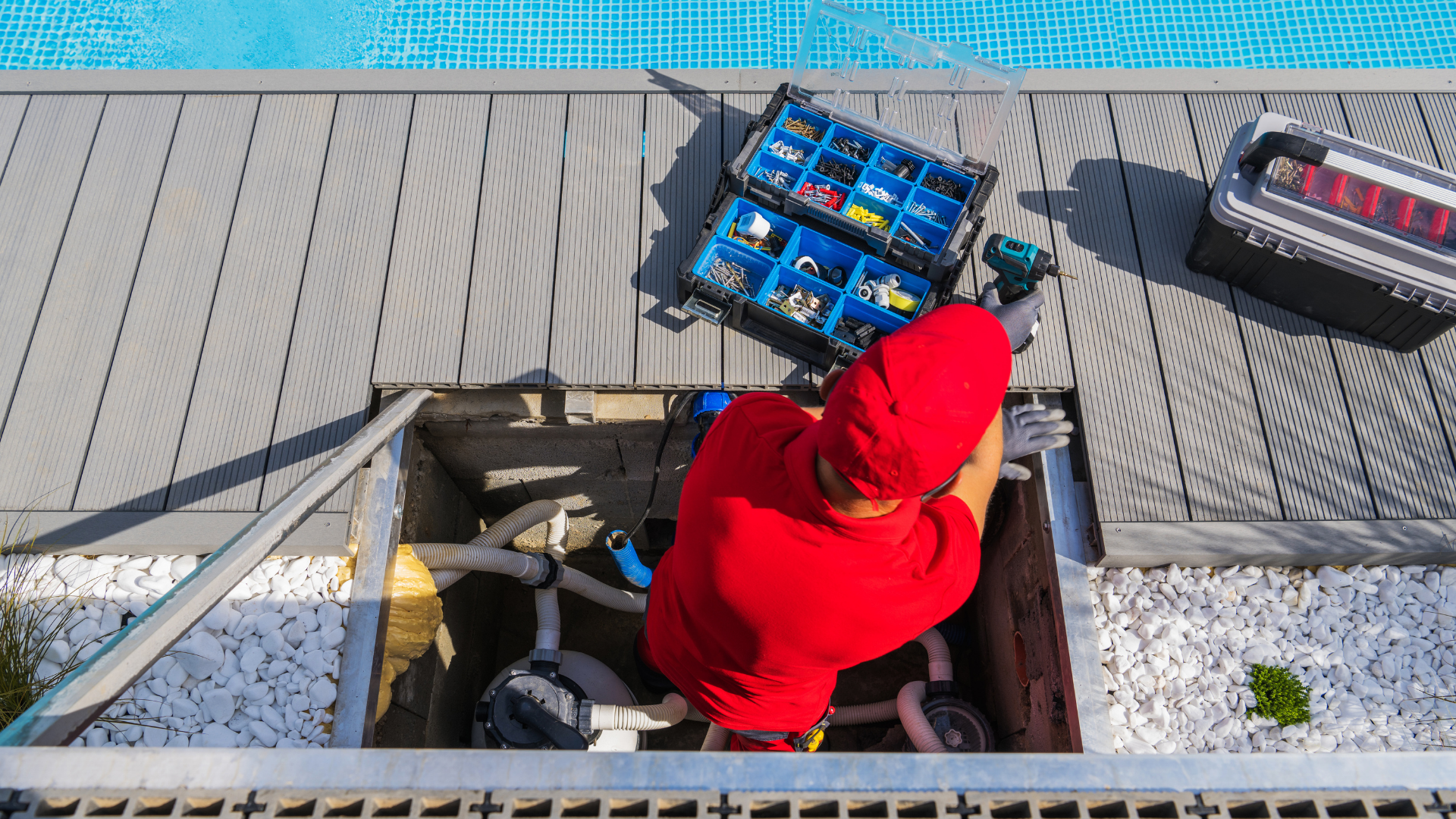 Person in a red hoodie stands in a rooftop hatch, next to blue crates and a black case.