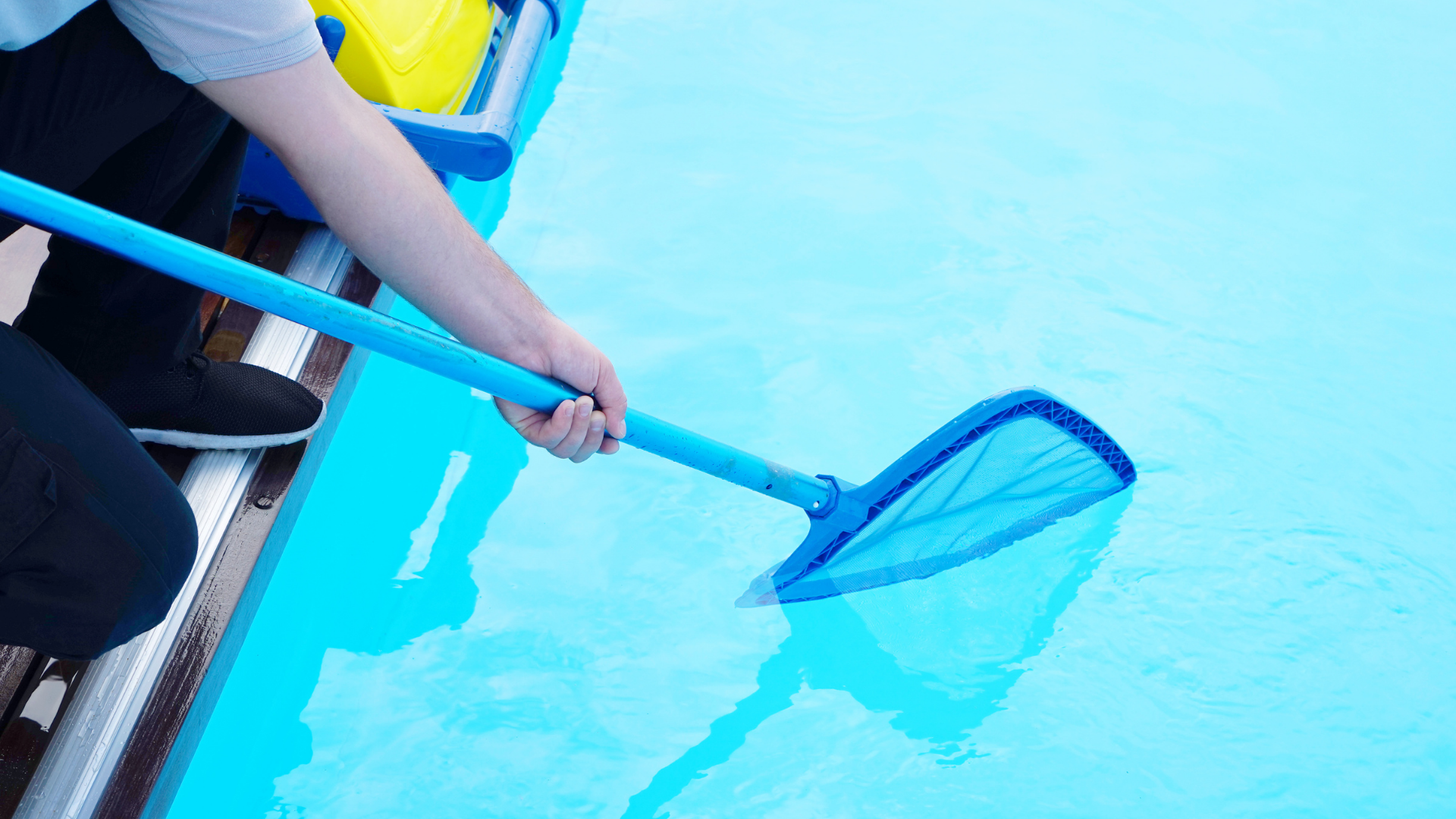 Person skimming debris from a bright blue swimming pool with a blue net pole