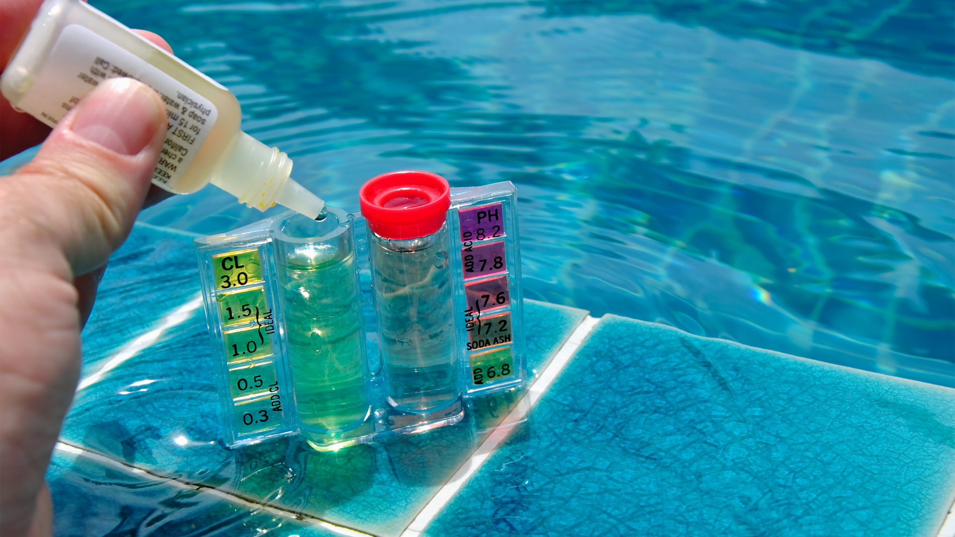 Hand pouring pool test liquid into color-coded water test vials beside a blue swimming pool.