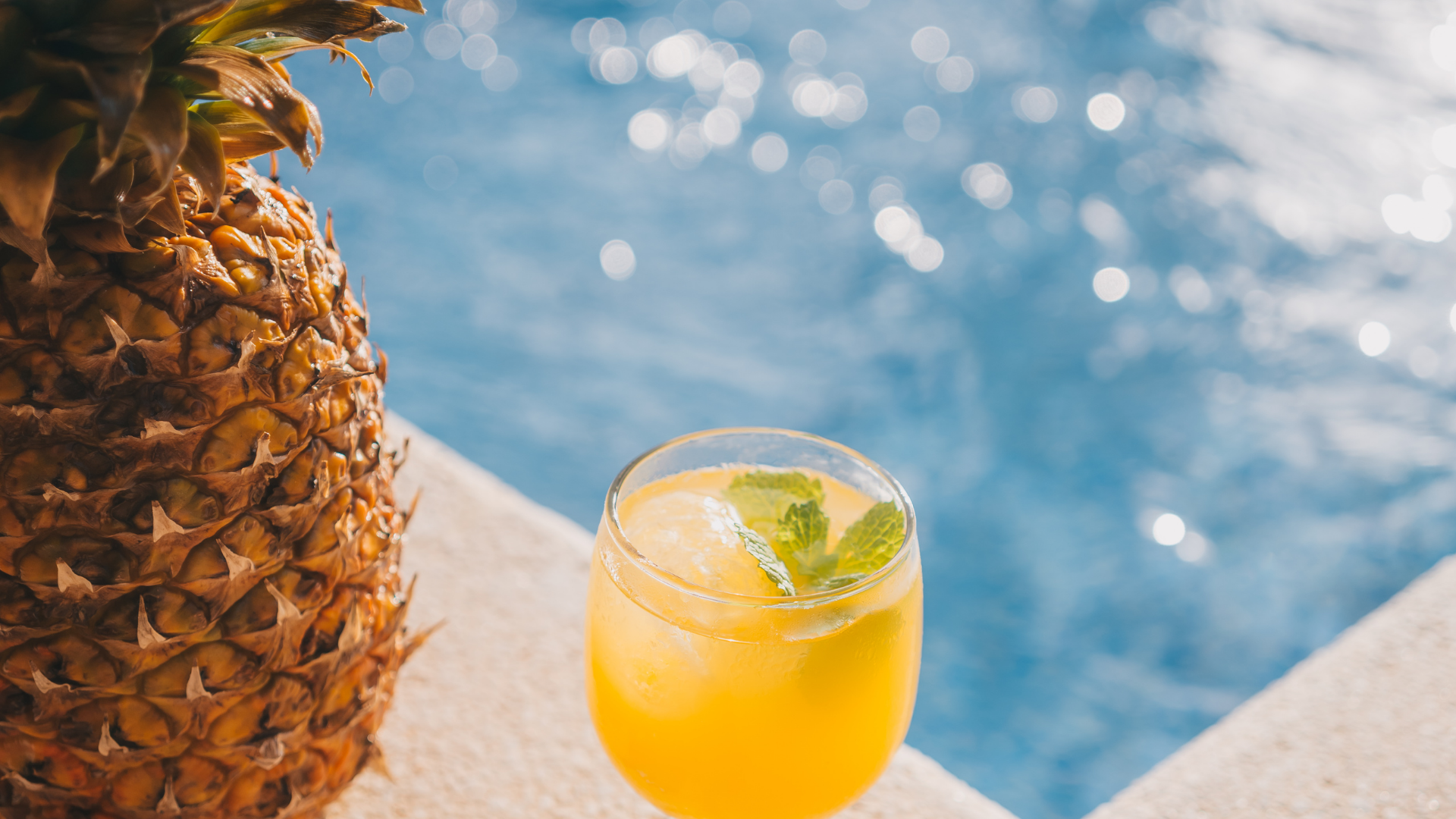 Tropical drink with mint beside a pineapple at a poolside with blue water