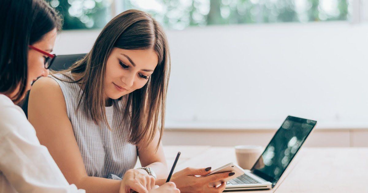 duas mulheres estão sentadas a uma mesa com um laptop e um telefone celular.