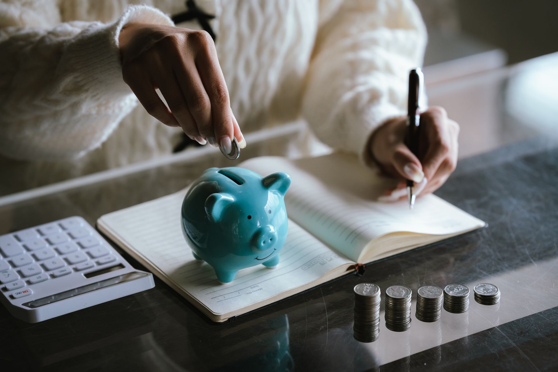 Blue piggy bank with stacked coins on a table, while a person uses a calculator in the background